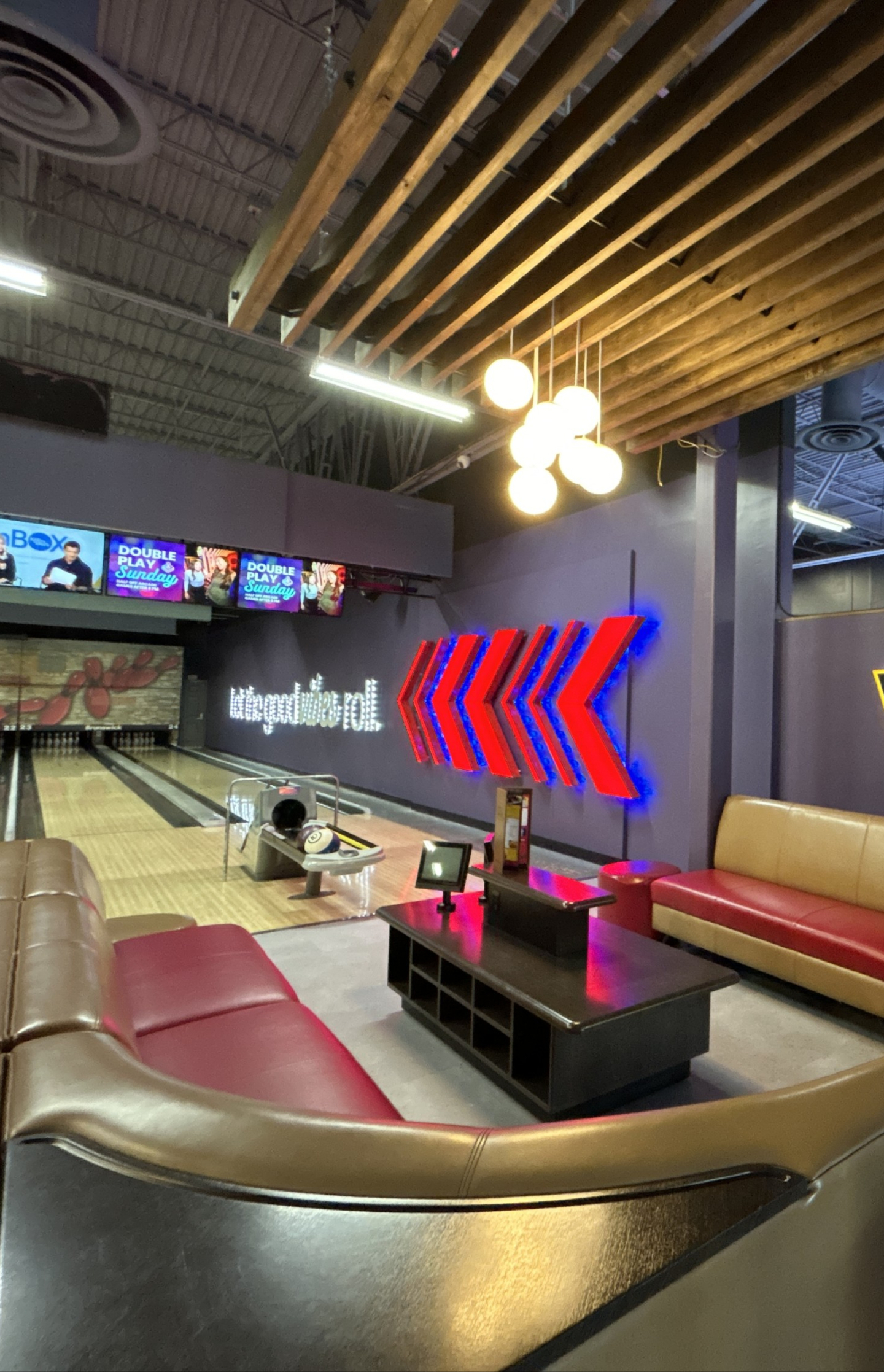 A VIP bowling alley seating area with leather booth seating and a low table beside illuminated bowling lanes. Wooden ceiling slats, hanging globe lights, and glowing lane graphics create a modern entertainment setting.